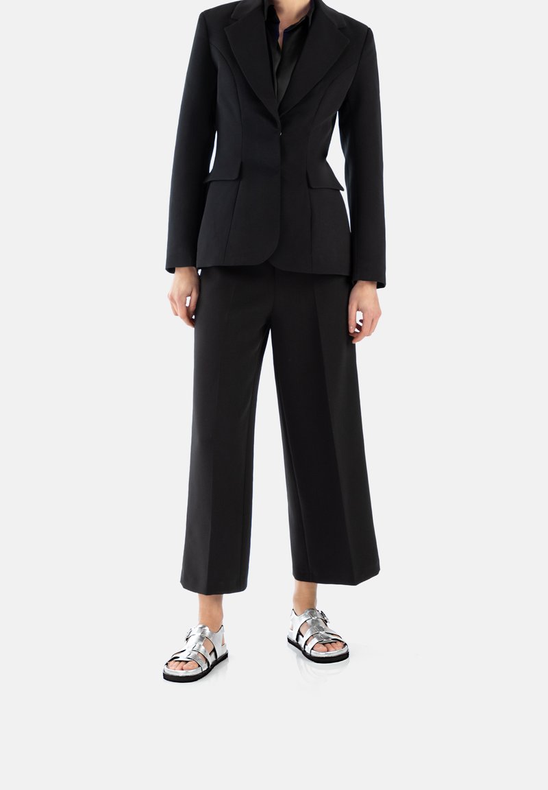 Blazer nero su misura con rever a picco, abbinato a pantaloni neri a gamba larga e sandali metallici argento con molteplici cinturini e suole nere.