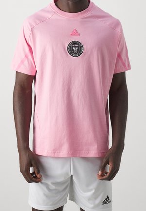 Mężczyzna w różowej koszulce Adidas z logo Inter Miami CF i białych szortach Adidas na jednolitym tle.