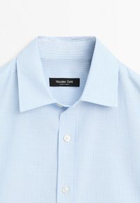 Jasnoniebieska koszula z cienkimi białymi pionowymi paskami, białymi guzikami i kołnierzykiem typu spread, rozłożona płasko, ukazująca metkę marki wewnątrz kołnierza.