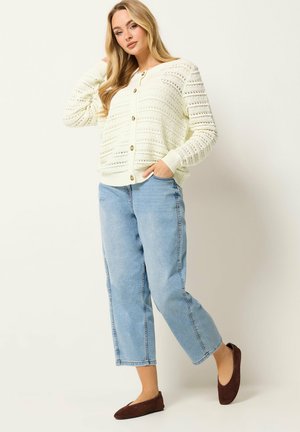 Junge Frau trägt einen weißen, gestrickten, geknöpften Pullover, hellblaue, verkürzte Jeans und braune flache Schuhe, steht vor einem schlichten Hintergrund.