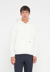 White fleece hoodie with a zipper and two front pockets. Features a soft texture and a small silver tag on the front.