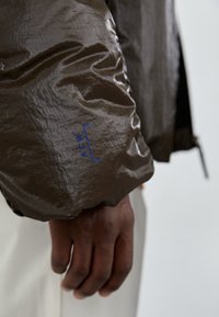 Brun, texturerad jacka med en glänsande finish, som har en blå "ACW" etikett på ärmen. Designen betonar en modern, minimalistisk stil.