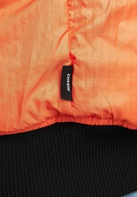 Oranje quilted stof met een klein zwart label met de tekst "CHASIN'" boven een brede zwart geribbelde boord.