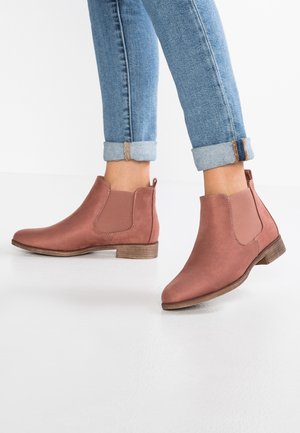 Brune Chelsea-støvler af ruskind til ankellængde, båret med opklemte blå jeans på en hvid baggrund.