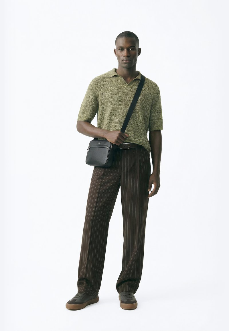 Man standing, wearing green textured short-sleeve polo, brown pinstripe pants, brown sneakers, and black crossbody bag.