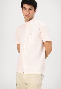 Camicia button-up a maniche corte di colore rosa chiaro, in tessuto testurizzato, con una tasca singola e un piccolo dettaglio con logo sul petto.