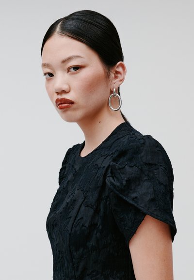 Schwarzes, strukturiertes Kleid mit kurzen Puffärmeln. Der Stoff weist ein erhabenes Muster auf. Das Modell trägt silberne Creolen und hat das Haar nach hinten gegelt.