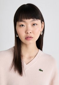 Pull léger rose en V fabriqué dans un tissu texturé, avec un petit logo de crocodile vert sur la poitrine. Fini lisse, coupe décontractée.