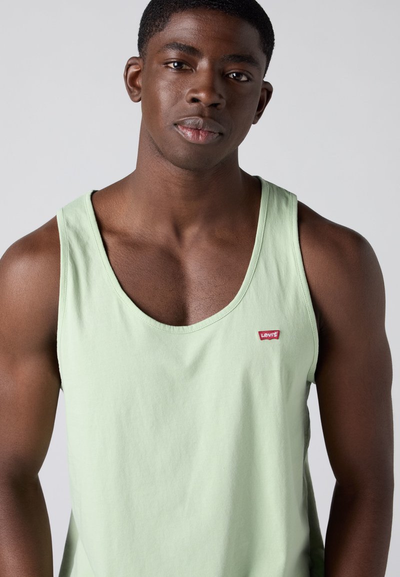 Jonget zwarte man met kort haar die een lichtgroene Levi’s tanktop draagt, kijkt naar voren tegen een effen grijze achtergrond.