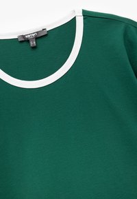 Camiseta de algodón verde con un cuello redondo acanalado blanco, que muestra una textura suave y un diseño casual; la etiqueta muestra la talla M y el nombre de la marca.