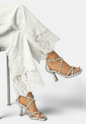 Voeten in zilveren sandalen met hoge hakken en strasstenen bandjes, gecombineerd met een witte broek met brede kanten rand, zittend met gekruiste benen.