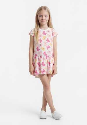 Fiatal lány kinyújtott lábakkal áll, fehér ruhát visel rózsaszín és sárga pillangós mintákkal, valamint fehér slip-on cipőben.