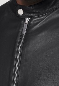 Black leather jacket with a smooth texture, featuring a high collar, silver zipper, and a snap button closure. Zipper detail is prominent.