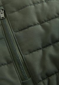 Gros plan d’un tissu de veste matelassée vert olive montrant une fermeture éclair en métal détaillée et des lignes de couture horizontales.