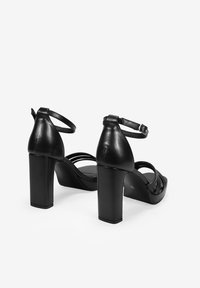 Sandalias de tacón negro de cuero con tacón grueso y tira en el tobillo. Presentan un diseño de tiras cruzadas y una textura suave. Estética minimalista.