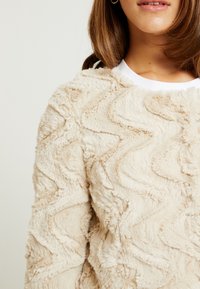 Beige pehmeä neule, jossa aaltoileva kuvio, teksturoitu pinta ja pyöreä pääntie; kerrostettu valkoisen paidan päälle.