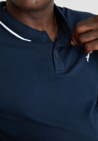 Tumedat sinise värviga polo särk valge triibulise kraega, millel on tekstuuriga kanga, kolmepunktiline nööpide rida ja tikandiga logo rinnal.