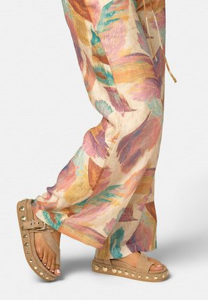 Persona con pantalones sueltos de estampado colorido y sandalias beige con plataforma y tachuelas metálicas, con los dedos pintados de rosa claro.