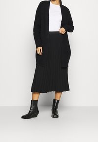 Cardigan negro texturizado sobre una camisa blanca, combinado con una falda negra plisada y botas negras de tobillo, mostrando un diseño de atuendo en capas.