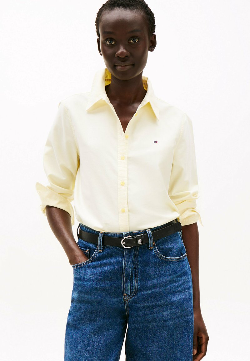 Camisa amarilla de botones con cuello, mangas enrolladas, combinada con jeans azul oscuro y un cinturón negro con una hebilla de plata.