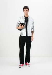 Joven de pie, vistiendo pantalones negros, una camisa negra, una camisa a rayas blancas, y zapatillas rojas, sosteniendo una chaqueta negra doblada frente a un fondo blanco.