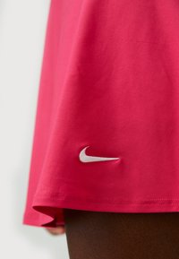 Fustă sport roz, realizată dintr-un material neted, cu un tiv curbat și un logo Nike brodat alb în partea stângă jos.