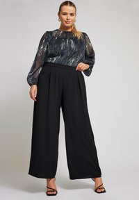 Durchscheinende, gemusterte Bluse in Dunkelblau mit langen, luftigen Ärmeln; hochtaillierte schwarze weit geschnittene Hose; schwarze Sandalen mit Riemen.