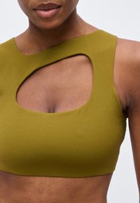 Kvinde iført olivengrøn ærmeløs cropped top med asymmetrisk udsnit over brystet, vist fra skuldre til mave.