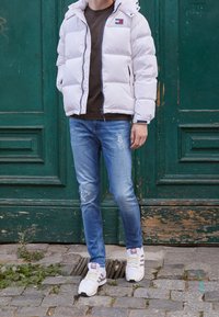 Hvid dunjakke med hætte, marineblå lynlås, båret over en mørk t-shirt. Blå slidte jeans og hvide sneakers med beige striber, stående på brosten.