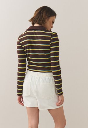 Woman facing away wearing a brown long-sleeve shirt with blue and yellow horizontal stripes and white elastic-waist shorts with back pockets.