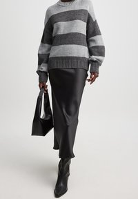 Henkilö, joka pukee päällensä harmaan ja tummanharmaan raidallisen neuleen, mustan nahkaisen midi-hameen, mustat saappaat ja pitää mustaa käsilaukkua valkoista taustaa vasten.