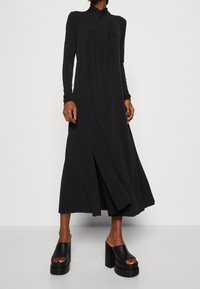 Mustassa pitkähihaisessa mekossa on korkea kaulus, kietaisumalli ja sivuhalkio. Sileä, valuva kangas ja rento istuvuus. Yhdistetty paksupohjaisiin mustiin sandaaleihin.