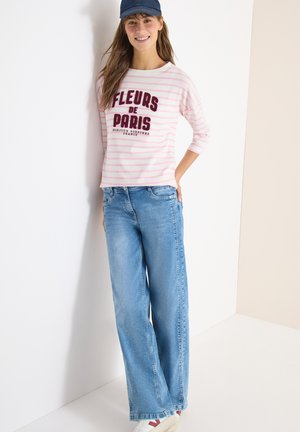 Kvinde iført en rosa og hvidstribet skjorte med teksten "Fleurs de Paris", blå viddebukser, hvide sneakers og en mørkeblå baseballkasket.