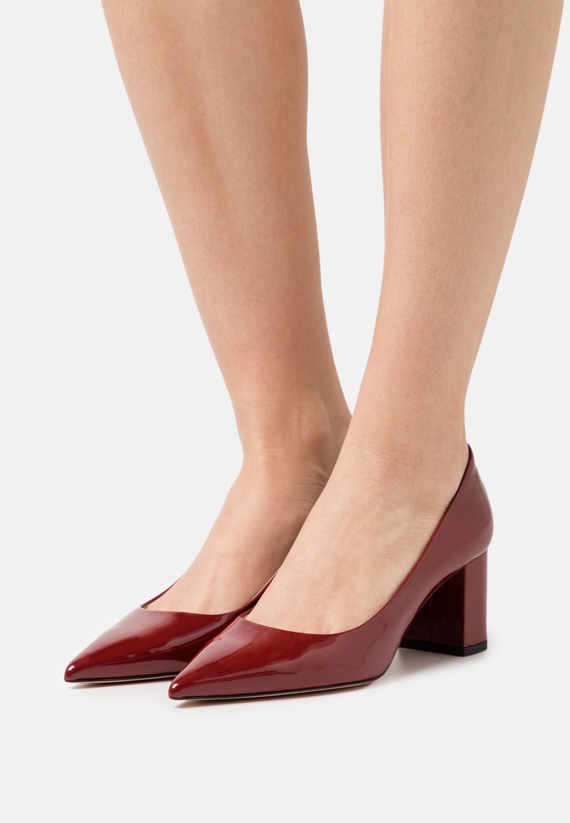Jambes portant des escarpins à talons bas blocs en cuir verni rouge foncé brillant, avec des bouts pointus, sur un fond clair.