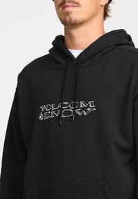 Hoodie noir avec cordons de serrage, arborant un logo "Volcom Snow" brodé en blanc. Texture douce, poche kangourou à l'avant.