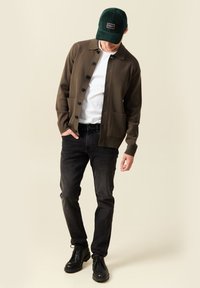 Hombre con una gorra verde oscuro, un cárdigan marrón con botones, una camiseta blanca, jeans negros y botas negras, de pie con la cabeza inclinada hacia abajo.