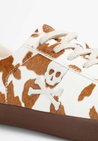 Zapatillas con un diseño de estampado de vaca en blanco y marrón, que cuentan con un detalle de calavera y tibias cruzadas, y una suela de goma marrón con agarre texturizado.