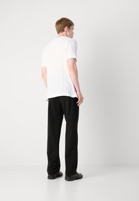 Cămașă albă texturată cu mânecă scurtă, vedere din spate, asociată cu pantaloni negri și pantofi, stând pe un fundal alb simplu.