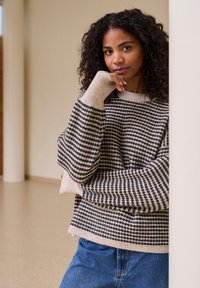 Pull rayé beige et gris foncé, texture tricotée, design oversize avec des manches larges, associé à un jean bleu, debout contre un mur.