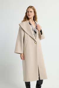 Abrigo beige con un cuello ancho y un único botón circular, que cuenta con bolsillos laterales y un tejido liso y estructurado. Mangas largas con un ajuste relajado.