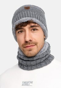 Mann trägt eine graue Strickmütze und einen passenden Halswärmer, gefüttert mit weichem Fleece, bekleidet mit einem weißen Hemd mit Text auf der Brust.