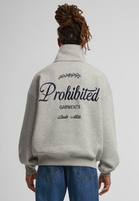 Sudadera gris de cuello alto, con el logo bordado de "Prendas Prohibidas" en la parte posterior y mangas puños. Acentos azules.