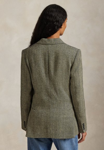 Vrouw draagt een grijze blazer met visgraatmotief en blauwe spijkerbroek, kijkt met lang donker haar van achteren tegen een effen achtergrond.
