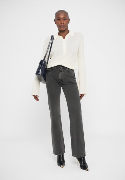 Persona de pie con un suéter de punto color crema, jeans gris oscuro, botas de tacón negras y llevando un bolso de hombro azul marino, todo sobre un fondo blanco.
