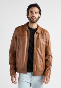 Man met krullend donker haar en een baard, gekleed in een bruine leren jas over een zwart shirt en lichtblauwe jeans, staande tegen een effen achtergrond.