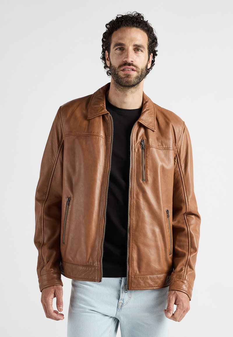 Man met krullend donker haar en een baard, gekleed in een bruine leren jas over een zwart shirt en lichtblauwe jeans, staande tegen een effen achtergrond.