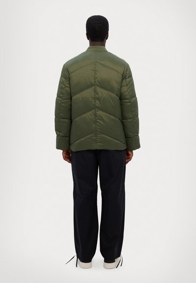 Veste matelassée vert olive avec un design en piquage, dotée d'un col haut et de poignets côtelés. Associée à un pantalon sombre et ample et à des baskets blanches.