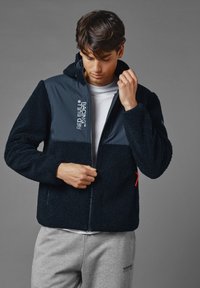 Zwarte fleecejas met marineblauwe schouders, kap en ritsluiting. Voorzien van Red Bull Racing branding en een rode ritssluiting aan de zijkant.