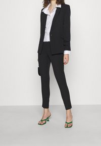 Mustat bleiseri, jossa on rakenteelliset olkapäät, valkoisen nappipaidan päällä, yhdistettynä viivallisiiin housuihin ja vihreisiin korkokenkiin.