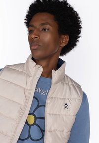 Joven con cabello rizado que lleva un chaleco acolchado beige sobre una sudadera azul con un gran gráfico de flor y texto.